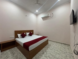A bedroom with a bed, a nightstand, a TV, and an air conditioning unit. - Hotel Baldev Palace (Vrindavan)