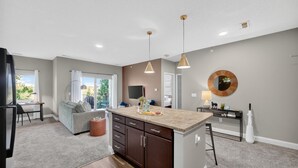 A modern kitchen with a central island, pendant lighting, and a dining area with a sofa and table. - Wooded Views | King Bed | No Stairs (West Des Moines)