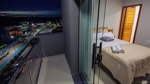 A balcony with a view of a cityscape at night, a bed with a white bedspread and pillows, and a wooden door. - Hotel Madalena - Beiro Rio (Sao Miguel Do Guama)