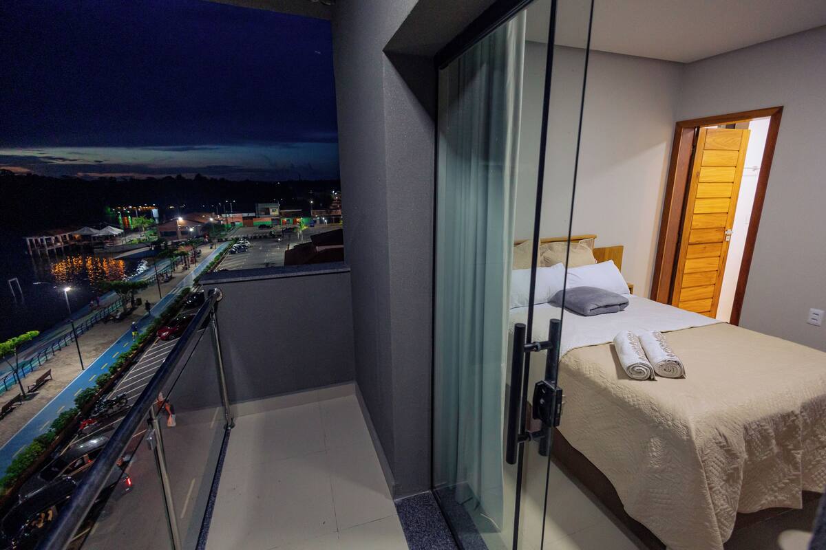 A balcony with a view of a cityscape at night, a bed with a white bedspread and pillows, and a wooden door.