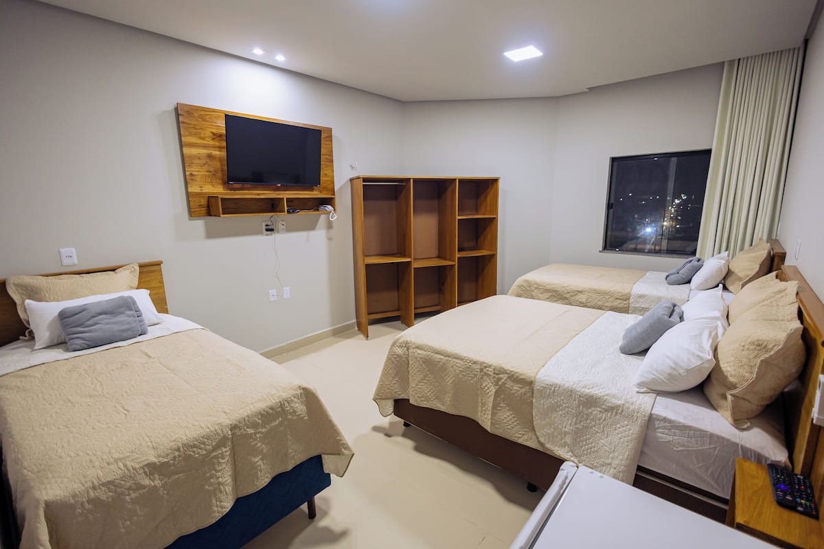 A hotel room with two beds, a TV mounted on the wall, a window with curtains, and a wooden shelf unit.