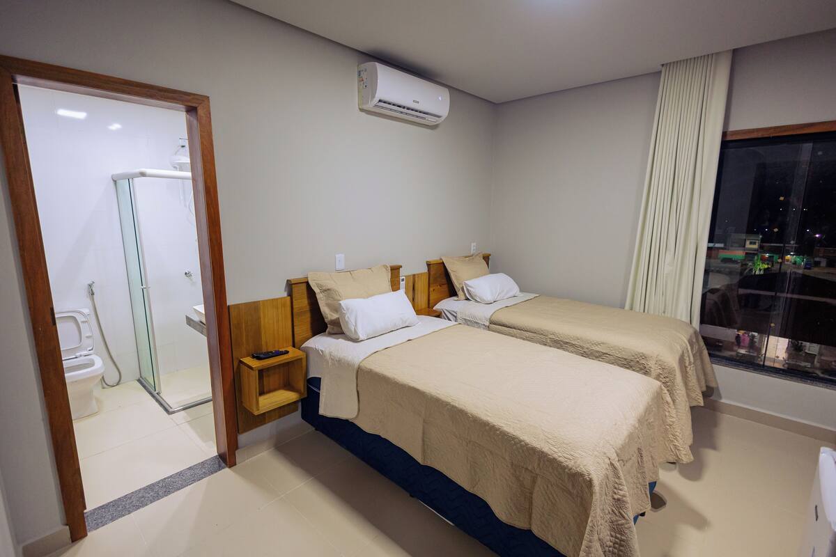 A hotel room with two beds, a shower, a window with a view, and an air conditioning unit.