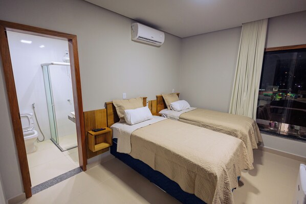 A hotel room with two beds, a shower, a window with a view, and an air conditioning unit.