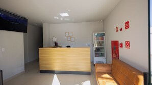 A reception area with a wooden desk, a couch, a refrigerator, and a TV on the wall. - Hotel Madalena - Beiro Rio (Sao Miguel Do Guama)