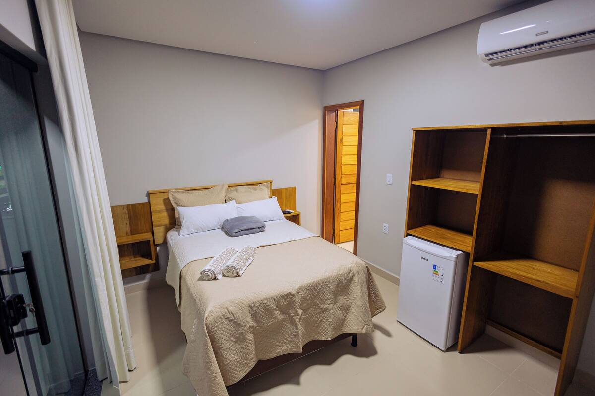 A bedroom with a bed, a small refrigerator, a wooden wardrobe, and a glass door leading to a bathroom.