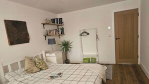 Un dormitorio con cama, mesita de noche, lámpara, planta en maceta, estantería, espejo y puerta de madera.