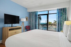 A hotel room with a large bed, a flat-screen TV, a wooden dresser, a desk with a lamp, and a balcony with a view of the ocean. - Pool View 1st Floor Room | King Bed | Kitchen | Dog Friendly (Myrtle Beach)