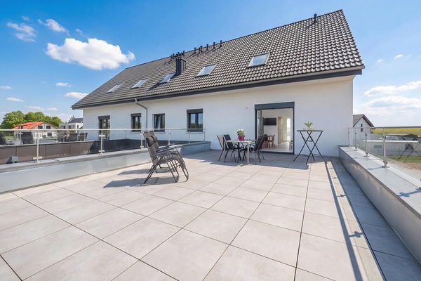 The exterior of the modern white building is displayed from the terrace, emphasizing its massive outdoor tiled living space. Glass railings run along the entire perimeter, offering panoramic views of the surrounding countryside.