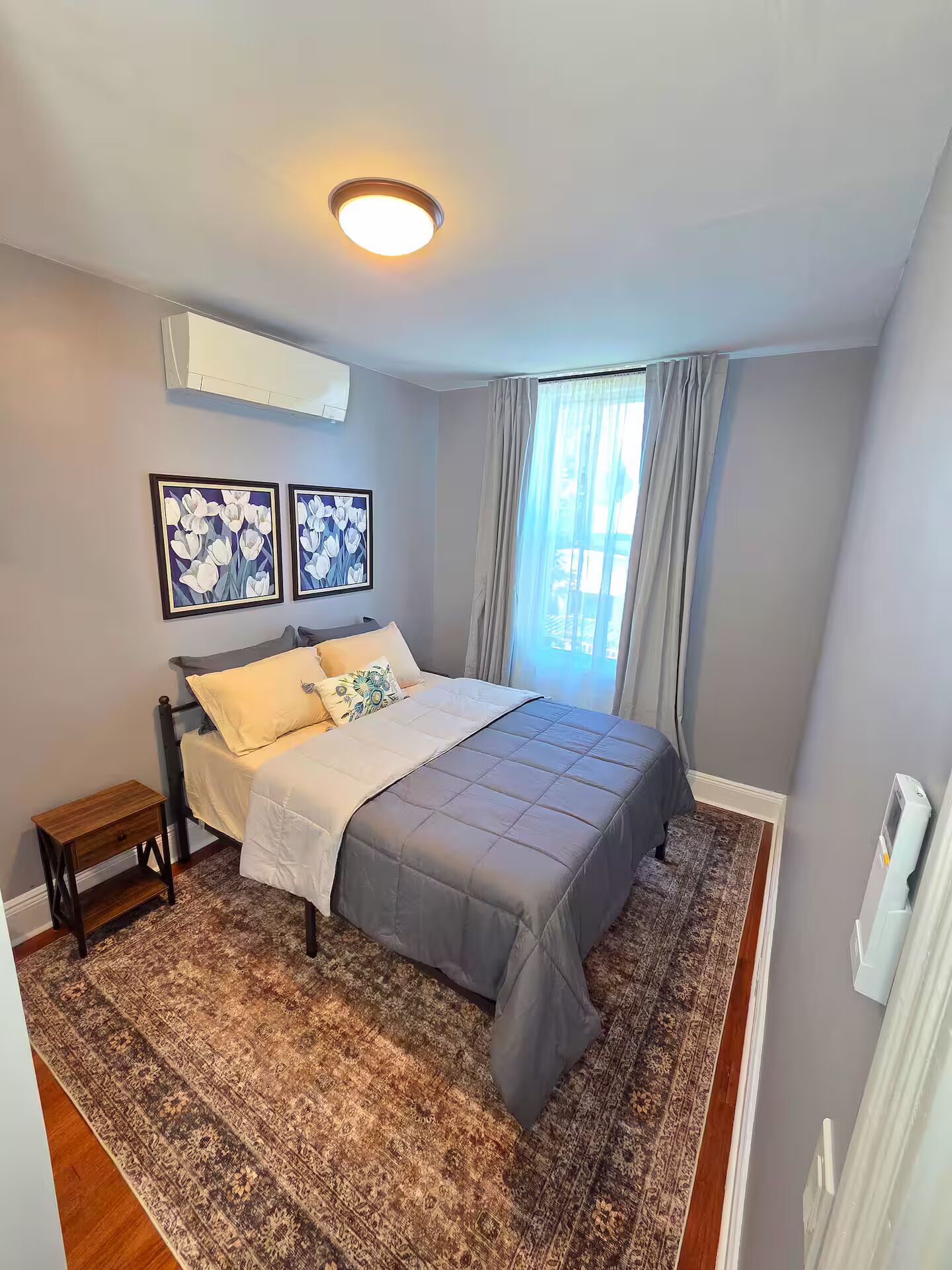 A bedroom with a bed, a small bedside table, a patterned rug, and two wall-mounted air conditioning units.