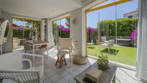 Una habitación amplia con un gran ventanal corredera que da a un jardín, un comedor con mesa y sillas, y una zona de estar acogedora con un banco acolchado y una mesita.