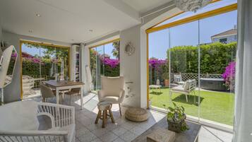 A spacious room with a large sliding glass door leading to a garden, a dining area with a table and chairs, and a cozy seating area with a cushioned bench and a small table.