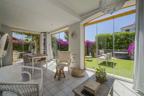 Ein geräumiges Zimmer mit einer großen Schiebeglasdoor zu einem Garten, einem Essbereich mit Tisch und Stühlen sowie einer gemütlichen Sitzecke mit gepolstertem Sitz und einem kleinen Tisch.