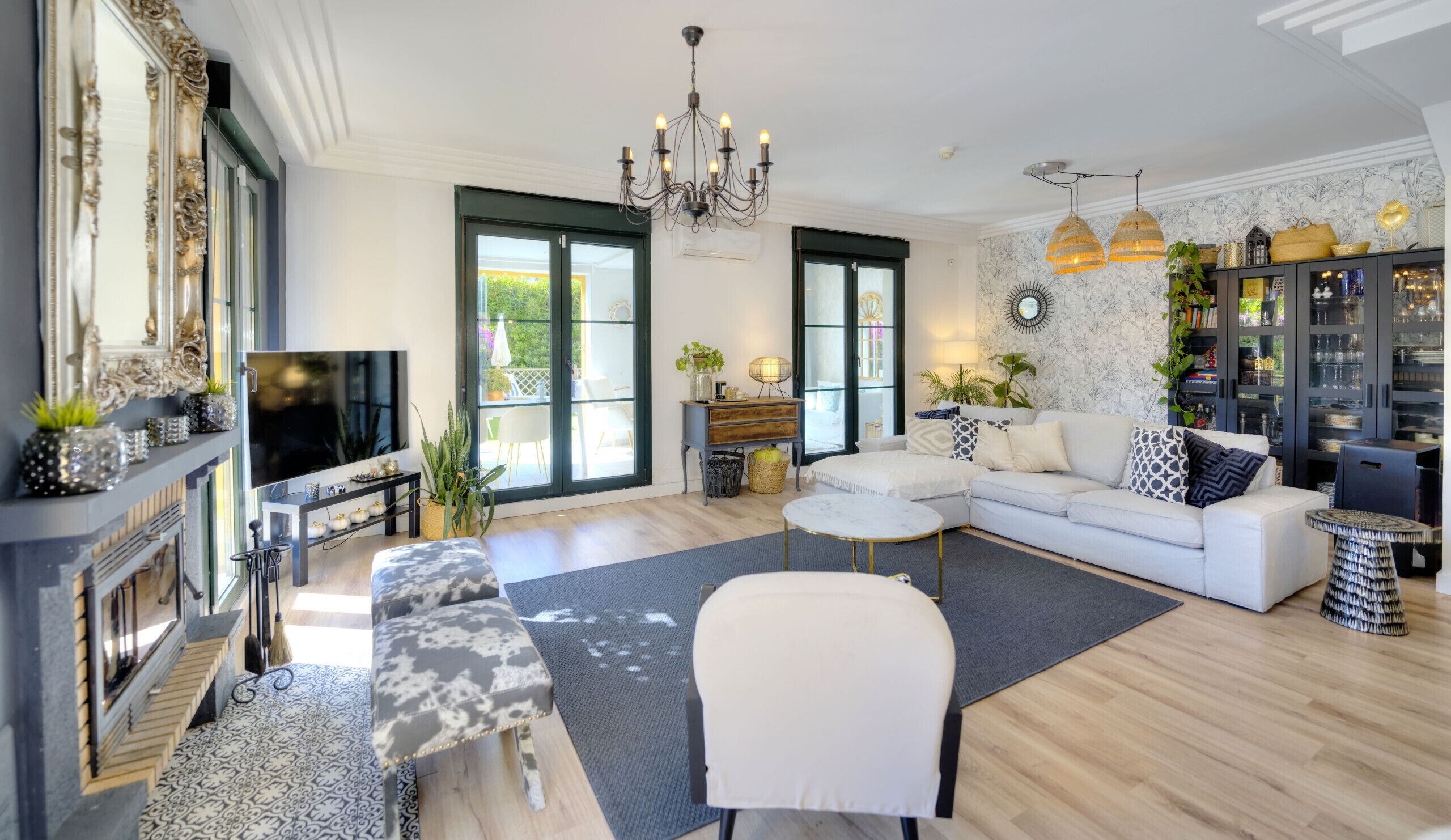 Un salon moderne avec un canapé, un pouf et un téléviseur. On y trouve une cheminée, un lustre et une grande fenêtre.