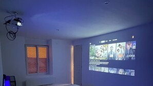 A bedroom with a bed, a wall-mounted projector displaying a show, and a window with blinds. - Spacious, newly renovated townhouse (Toul)