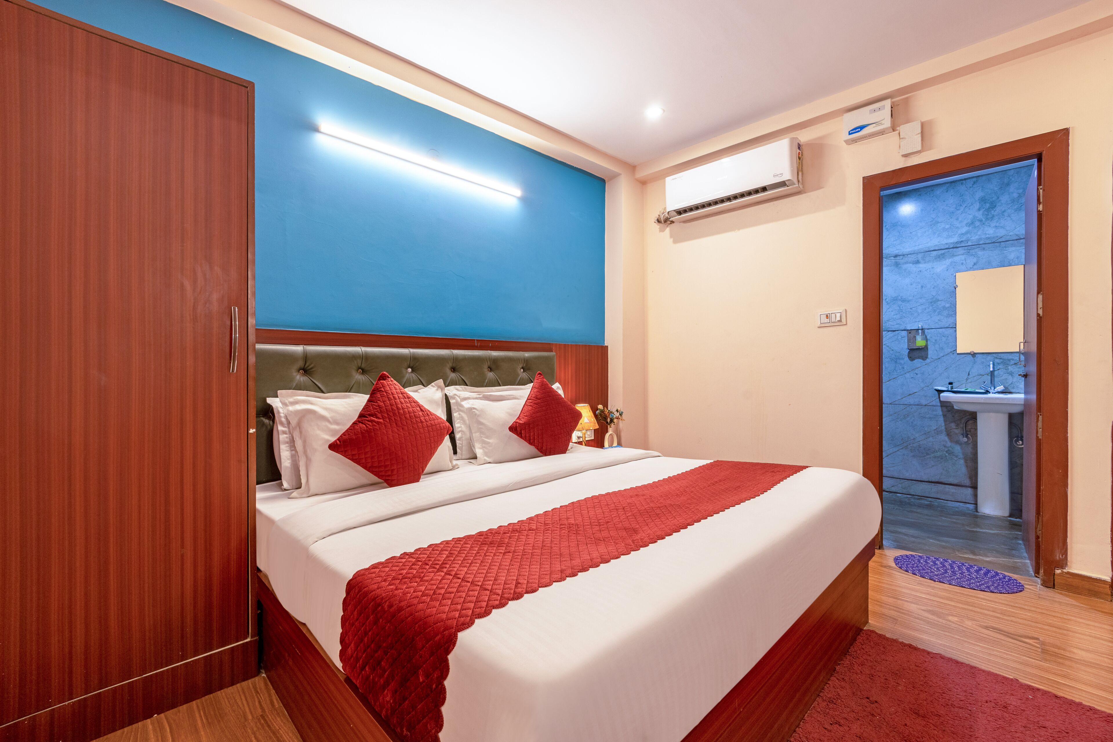 Habitación de hotel con una cama grande, un armario de madera, una pared azul decorativa, un equipo de aire acondicionado y un baño visible a través de una puerta abierta.