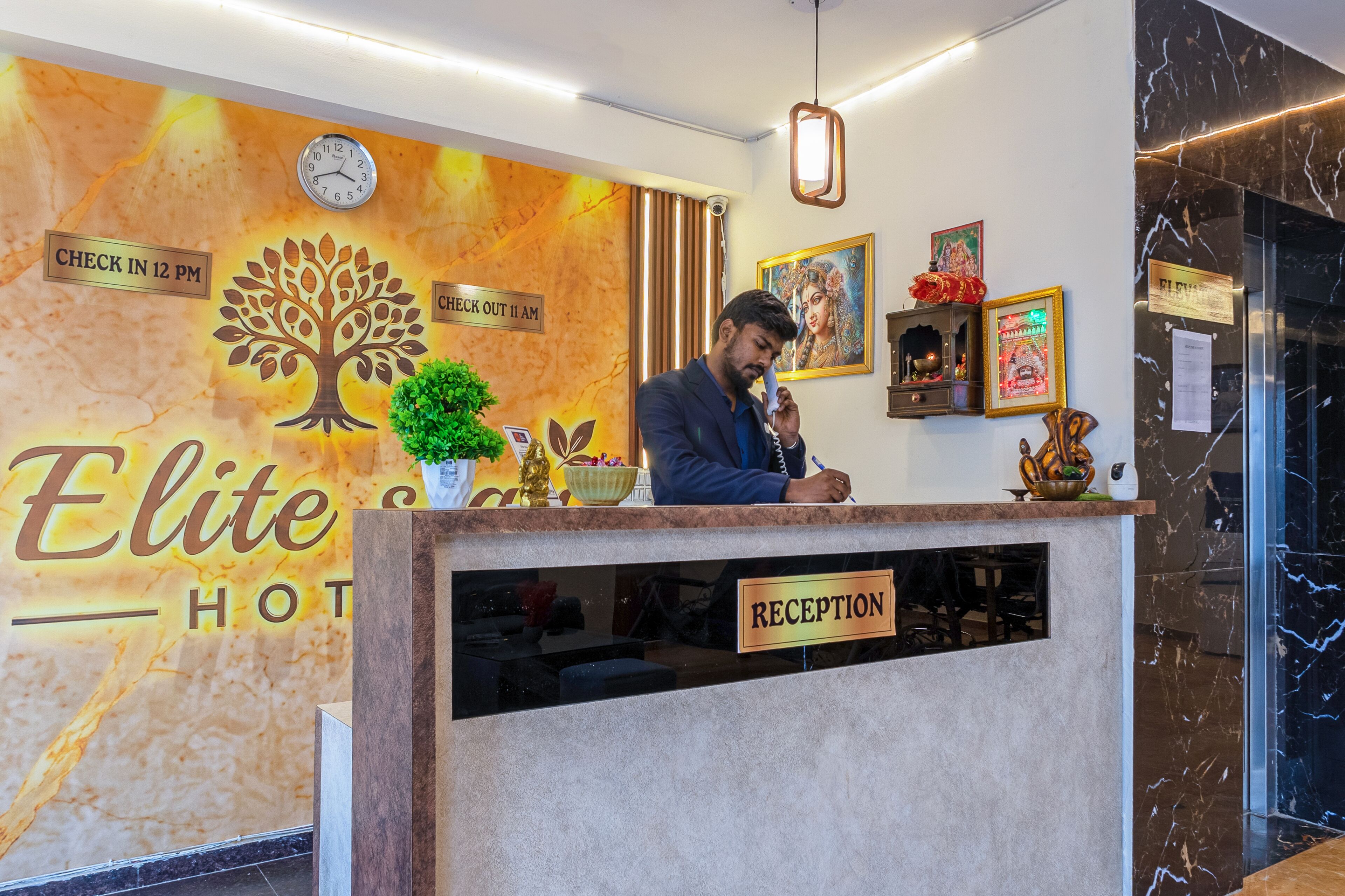 A hotel reception desk with a clock, a reception sign, and a wall with a tree and hotel name.