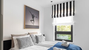 Una camera da letto con due letti, un'ampia finestra vista su del verde, una tenda a righe bianche e nere e un quadro appeso al muro.