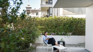 Una persona seduta su una sedia bianca in un patio, con un tavolino, circondata dal verde e un edificio sullo sfondo.
