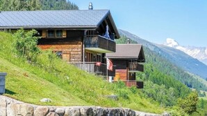 A wooden cabin with a balcony, set on a hillside with a mountain backdrop. - 4* renovated apartment at the top of the mountain at 1500 m (Goms)