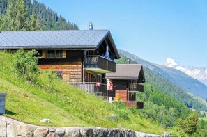 A wooden cabin with a balcony, set on a hillside with a mountain backdrop. - 4* renovated apartment at the top of the mountain at 1500 m (Goms)