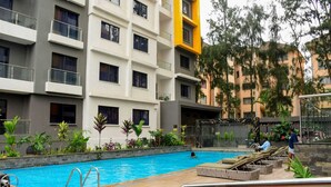 A swimming pool with lounge chairs and a wooden deck, surrounded by greenery and a multi-story building with balconies. - Clean, cozy and welcoming space for all my guests. (Nairobi)