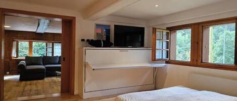 A bedroom with a bed, a TV on a shelf, a sofa in the adjacent room, and large windows.