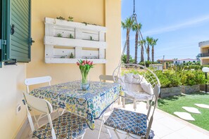 Eine Terrasse mit Tisch und Stühlen, einem Hängestuhl, einem weißen Pflanzgefäß mit Pflanzen und einem weißen, an der Wand befestigten Regal mit Pflanzen.