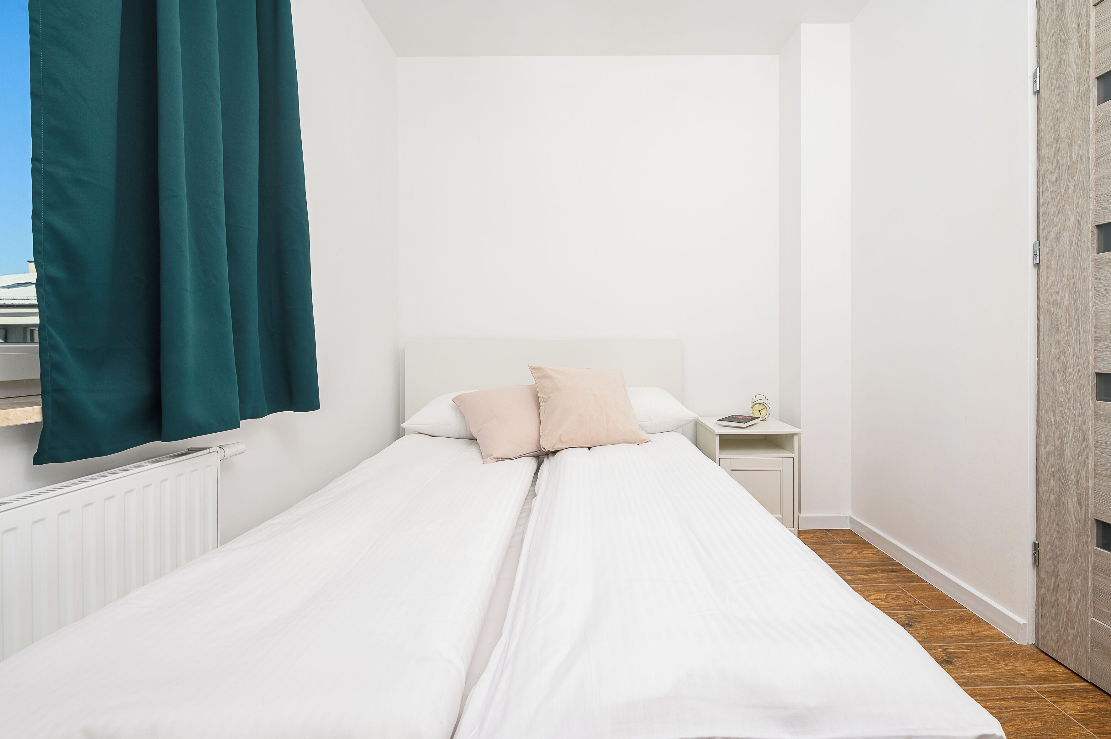 A single bed with white bedding, two pillows, a nightstand with a lamp, and a wooden door.