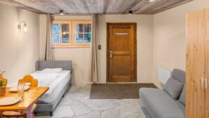 A room with a bed, a sofa, a wooden wardrobe, a dining table with chairs, a window with curtains, and a stone floor. - Cozy studio in the heart of an Alpine chalet (Anniviers)
