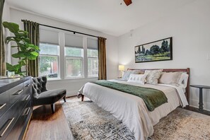 Ein Schlafzimmer mit einem Bett, einem Schminktisch, einem Sessel, einem Fenster mit Vorhängen, einem Bild an der Wand und einer Lampe.