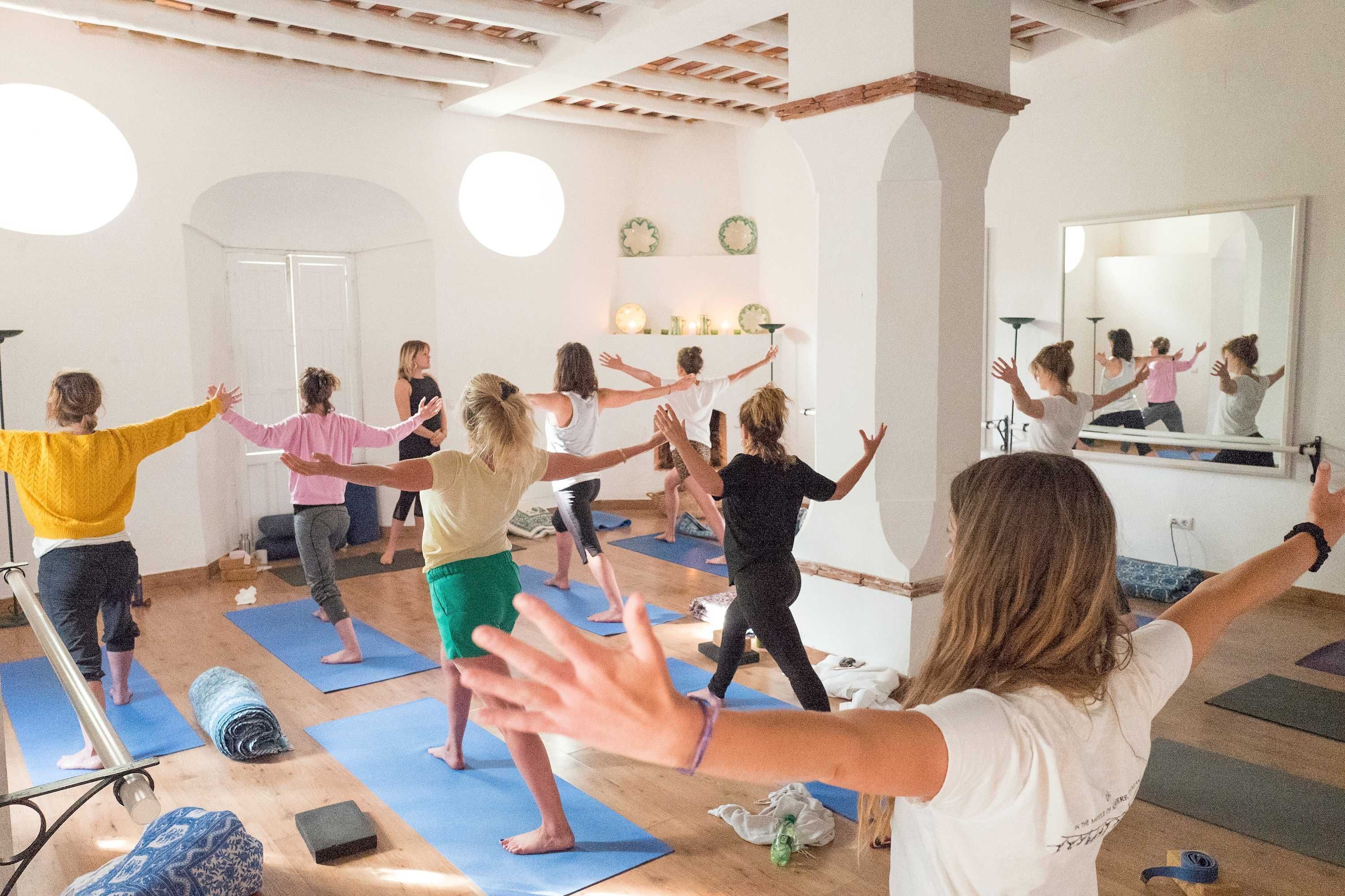 A yoga class in a room with wooden beams, a large mirror, and a white wall with shelves.