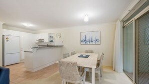 A compact kitchen with white cabinets, a built-in oven, and a small island with a sink. There is a dining area with a table and wicker chairs, and a painting of sailboats on the wall. - Westport Palms unit 4 (Port Macquarie)