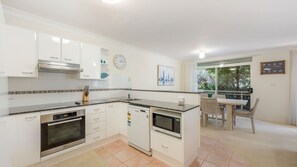 A modern kitchen with white cabinets, a built-in oven, a microwave, and a dishwasher. There is a dining area with a table and chairs, and a view of greenery through a window. - Westport Palms unit 4 (Port Macquarie)