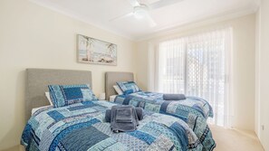 A bedroom with two beds, a ceiling fan, a large window with sheer curtains, and a beach-themed painting on the wall. - Westport Palms unit 4 (Port Macquarie)