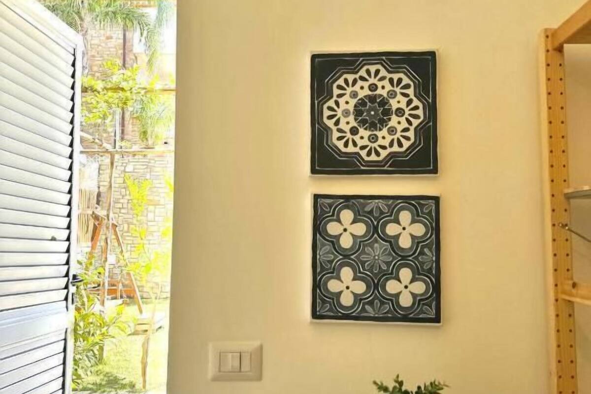 Una habitación con un azulejo decorativo en la pared, una mesita con una planta y una puerta que da a un espacio al aire libre.