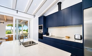 A modern kitchen with dark blue cabinets, stainless steel appliances, and a large window overlooking a scenic outdoor area. - Villa Irenal Javea, with Air Conditioning and Private Pool (Javea)