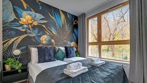 A bedroom with a large window, a bed with a dark quilt, multiple pillows, a bedside table with a lamp, and a wall mural. - Młoda Polska Apartments with parking  (Sopot)