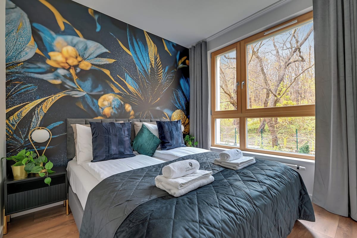 Un dormitorio con un ventanal grande, una cama con colcha oscura, varios almohadones, una mesita de noche con lámpara y un mural en la pared.