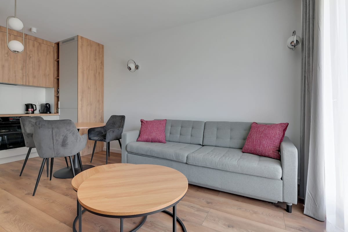 Una sala de estar moderna con un sofá gris, dos sillones grises, una mesa de centro de madera y una cocina con armarios de madera y una cafetera.