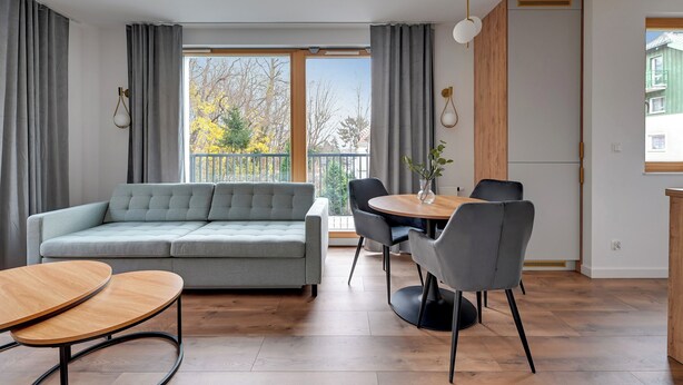A modern living room with a sofa, dining table, chairs, and a view through large windows. - Młoda Polska Apartments with parking  (Sopot)