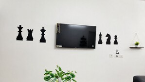 A room with a TV mounted on the wall, a black sofa, a small shelf with a plant, and a wall decal of chess pieces. - Cozy 2 & 1 Bedroom Apartment in Sahibzada Ajit Singh Nagar (Mohali)