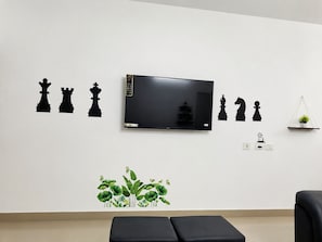 A room with a flat-screen TV mounted on the wall, a black sofa, a small shelf with a plant, and a wall decal of chess pieces.