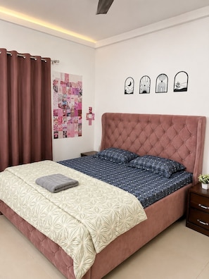 A bedroom with a pink bed, patterned bedding, a nightstand, a wall art collage, and a ceiling light.