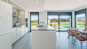 Une cuisine moderne dotée d’un îlot central, de grandes fenêtres et offrant une vue sur une piscine et un paysage verdoyant.