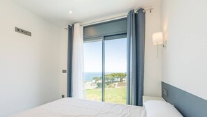 Une chambre à coucher avec un lit, une fenêtre avec des rideaux, un luminaire fixé au mur, et une vue sur la verdure et les bâtiments.
