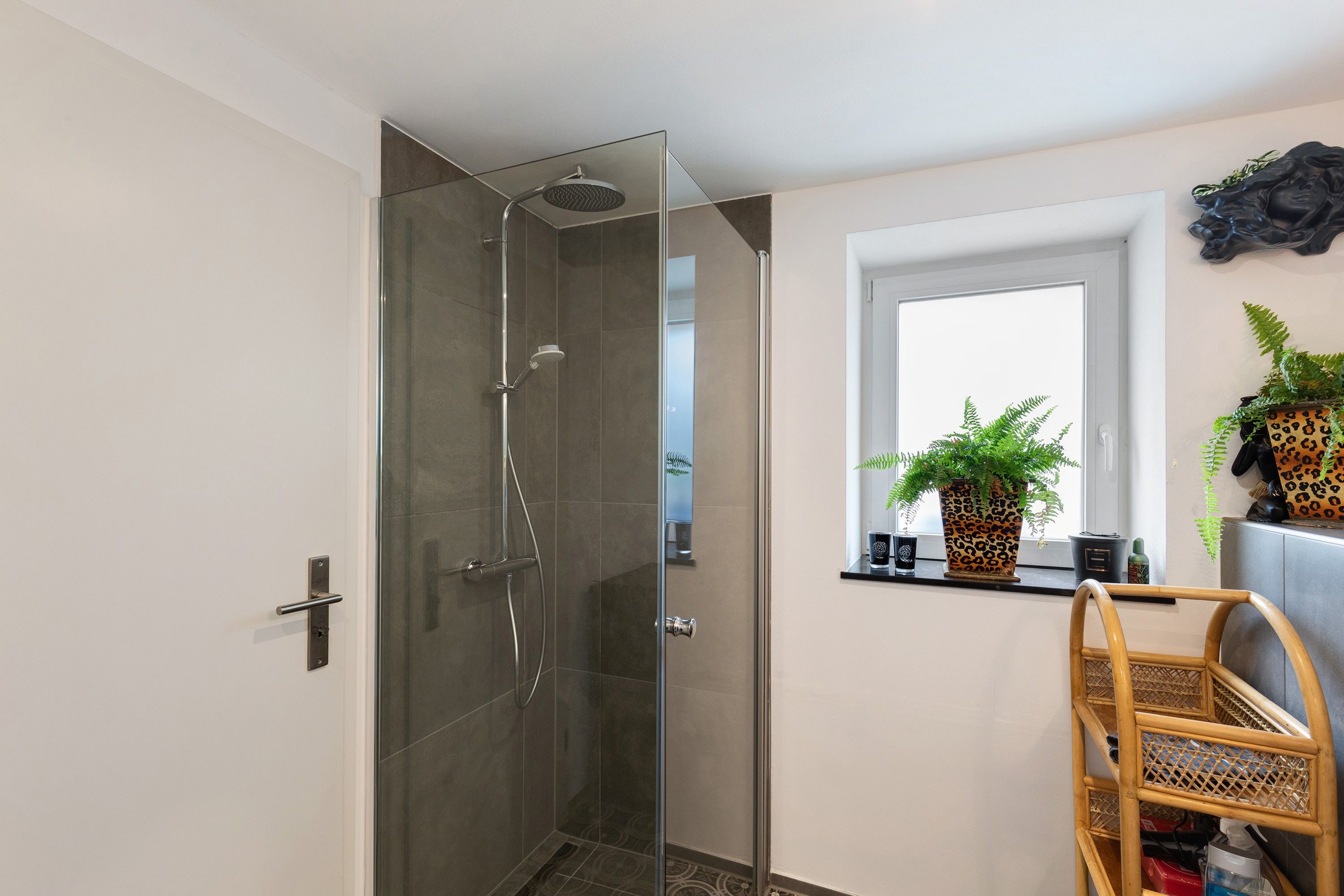 A modern bathroom with a glass-enclosed shower, a window with plants, and a wooden chair.