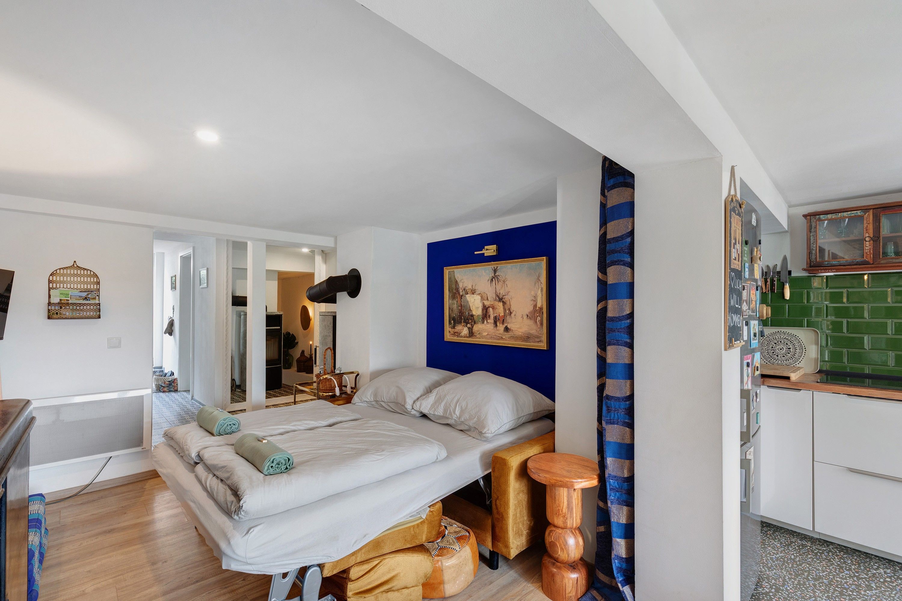 Quarto compacto com cama, uma mesa pequena, uma cadeira, um quadro na parede e um espaço com cozinha básica.