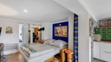 Quarto compacto com cama, uma mesa pequena, uma cadeira, um quadro na parede e um espaço com cozinha básica.