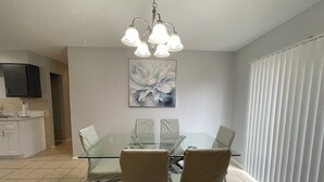 Et spiseområde med et glasbord, fire stole, en lysekrone, et stort blomsterbillede og udsigt til et hvidt gardin.
