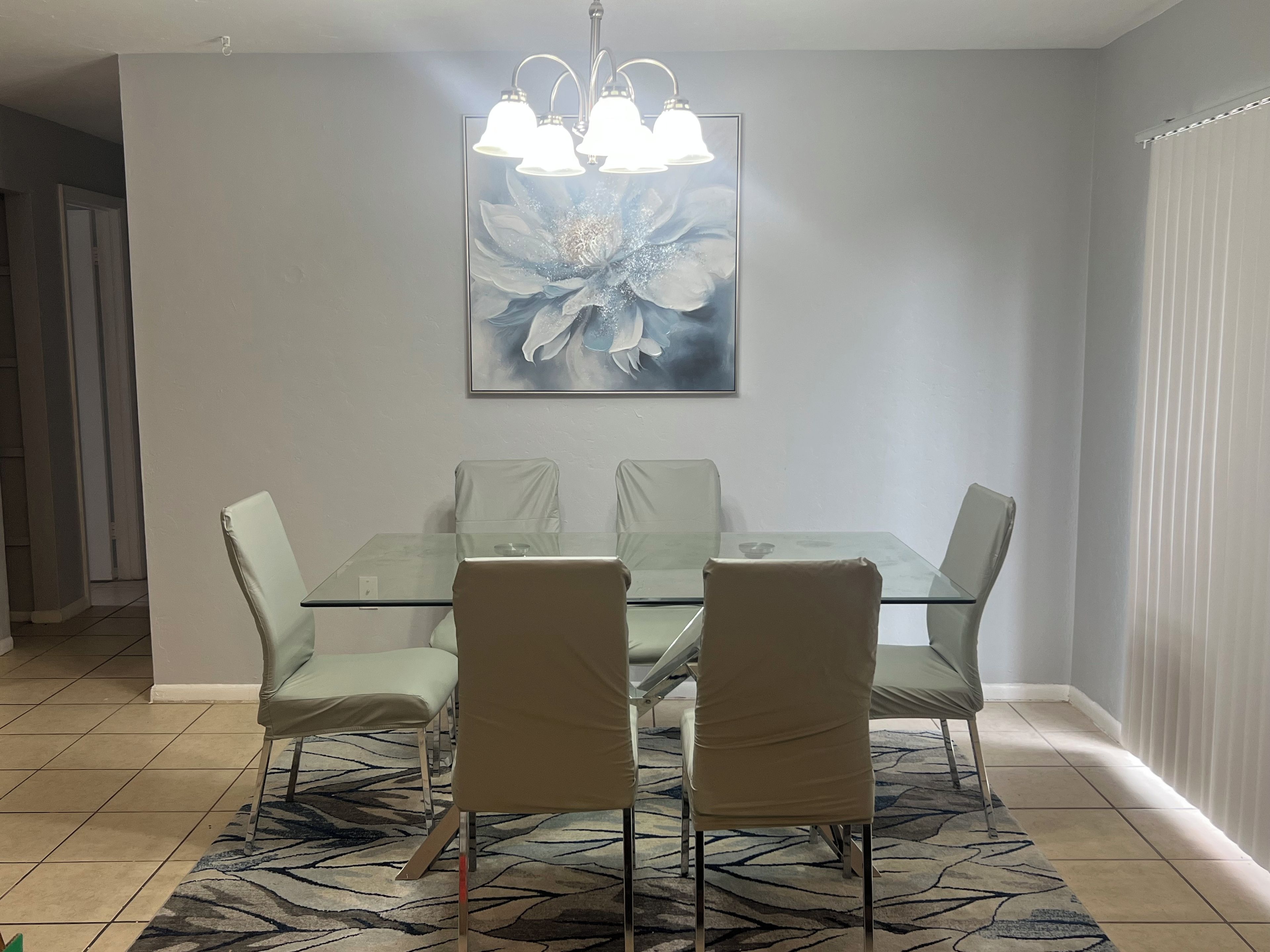 A dining area with a glass table and four chairs, a large floral painting on the wall, and a patterned rug on the floor.